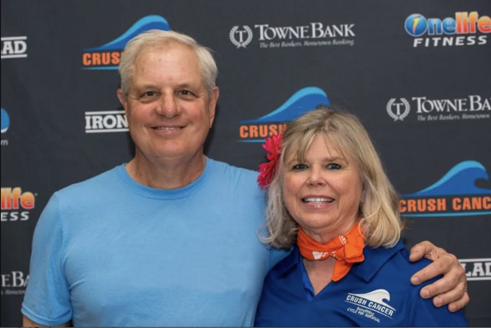 Tony Nero in a Cycle for Survival Virgina Beach shirt and Rosemary Wilson in a Crush Cancer shirt stand in front of a sponsor backdrop. 