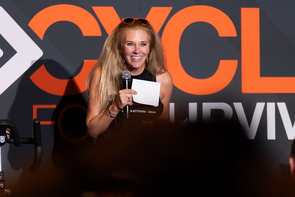 JJ Sailer speaks into a mic while holding paper cards in front of a Cycle for Survival backdrop. 