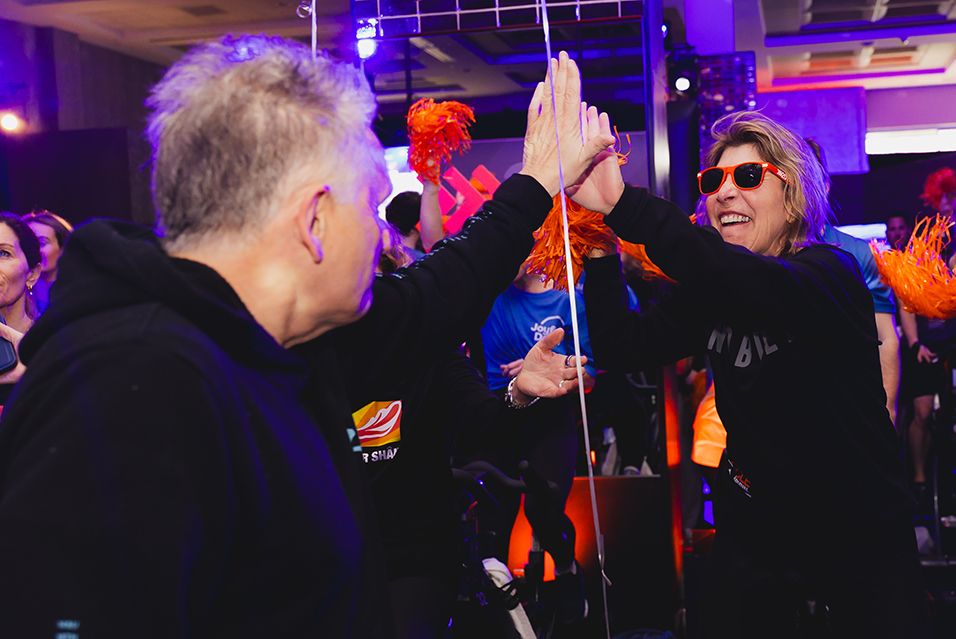 Two people high-fiving at an energetic cycling event, with vibrant lights and pom-poms.