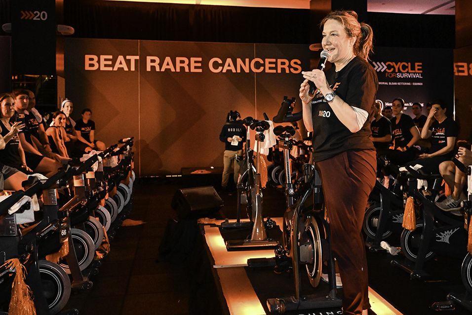 Jennifer James speaks to stationary-bike riders at a Cycle for Survival event. A banner reading “Beat Rare Cancers” hangs behind them. 