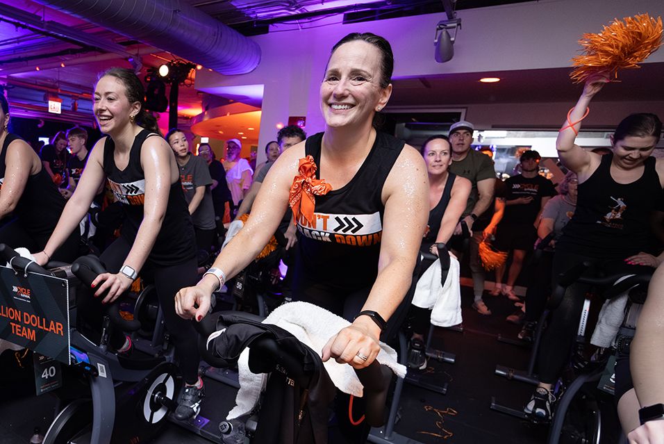 Julie Simon and other participants, wearing tank tops that read “DON’T BACK DOWN,” ride at a Cycle for Survival fundraising event. 