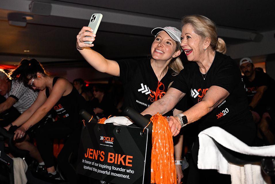 Kaitlin Lang takes a selfie with a fellow participant at a Cycle for Survival event. A sign on the rider’s bike reads “Jen’s Bike, in honor of our inspiring founder who sparked this powerful movement.”  