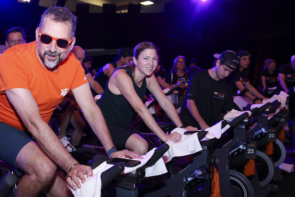 People cycling indoors with focused expressions under purple lighting.