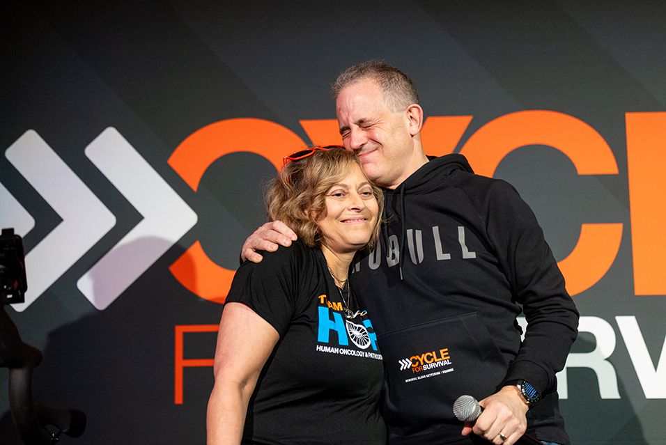 Dr. Ross Levine, wearing a NOBULL Cycle for Survival sweatshirt, and Marcy Kandel, wearing a Team HOPP T-shirt, embrace in front of a Cycle for Survival sign. 