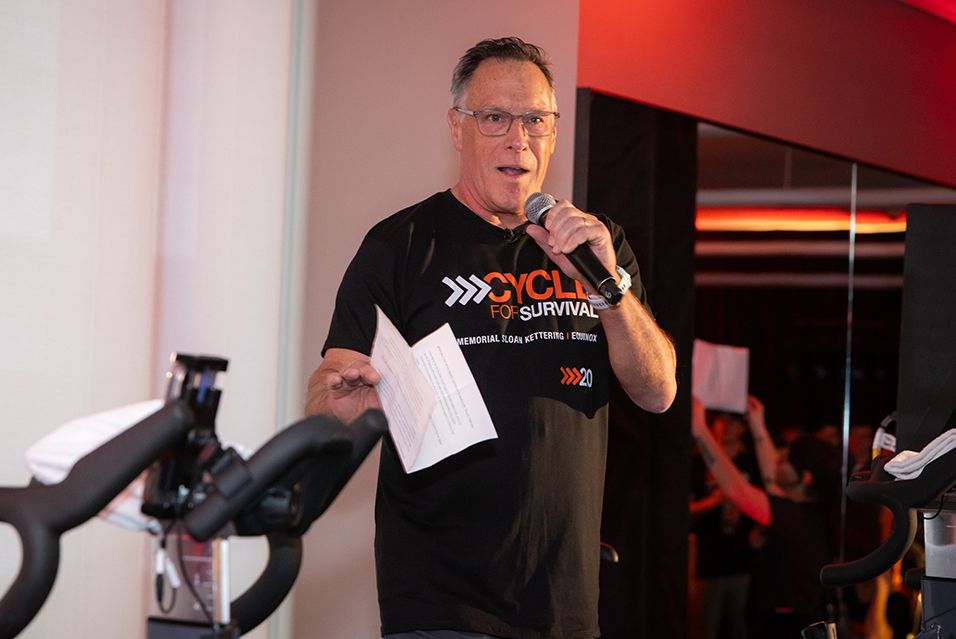 Man speaking into a microphone, holding papers, in a gym setting.