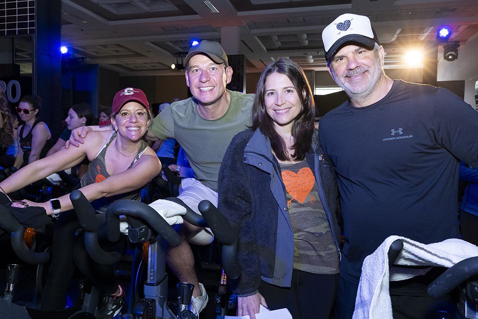 Four people smiling on stationary bikes at an indoor cycling event.