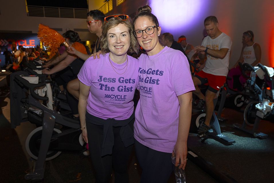 Jess and another participant wearing “Jess Gets The Gist” shirts at an indoor cycling event, with riders in the background.