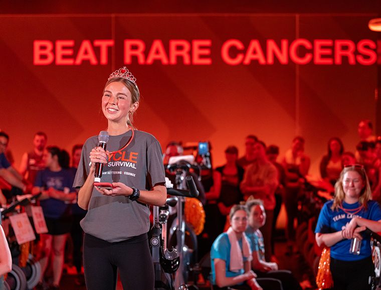 A Cycle for Survival participant wearing exercise clothes and a tiara stands at a mic, smiling and holding a cellphone, to address fellow riders.