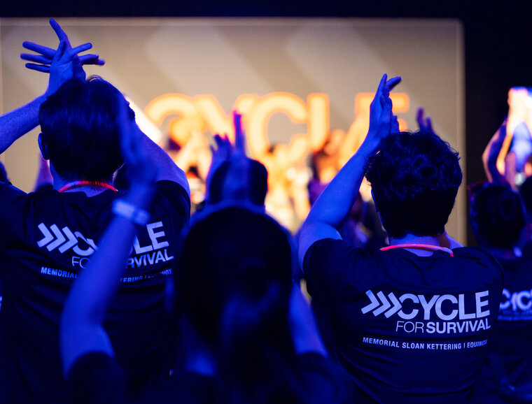 People in Cycle for Survival shirts clapping in a dimly lit room.