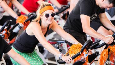 A woman wearing an orange headband, black tank top, green shorts, and white sunglasses sits on a stationary bicycle and looks at the camera.