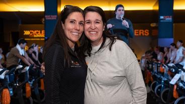 Two dark-haired women, one in a black sweatshirt, the other, a light brown top, stand smiling in front of people riding stationary bikes.