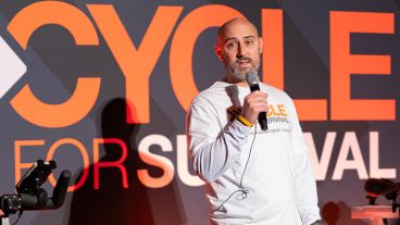 A man with a beard stands, wearing a Cycle for Survival shirt and holding a microphone.