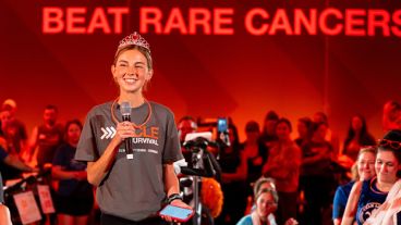 Cycle for Survival participant Sydney Towle, wearing a tiara and holding a mic, addresses riders beneath a “Beat Rare Cancers” sign. 