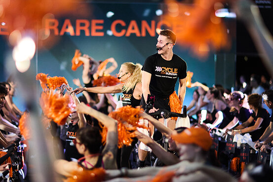 A man and a woman wearing a Cycle for Survival t-shirts smiles on stationary bikes surrounded by others riding stationary bikes and waving orange pom-poms. 
