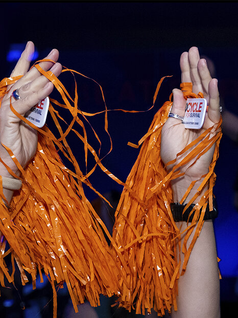 Two hands raised in the air hold orange pom-poms and tags that read “Cycle for Survival.” 