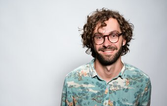 Man with curly hair, glasses, and a floral shirt smiling against a light background.