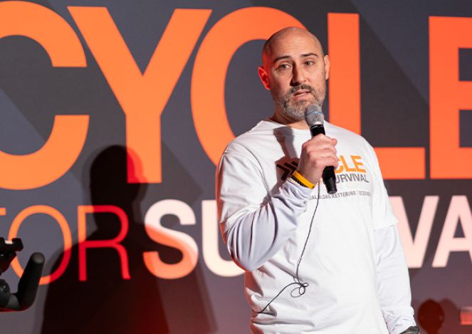 A man with a beard stands, wearing a Cycle for Survival shirt and holding a microphone.